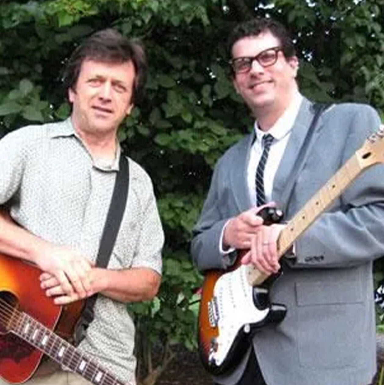 Ed Marra posing with a bandmate. One is holding an acoustic guitar, the other is holding an electric guitar